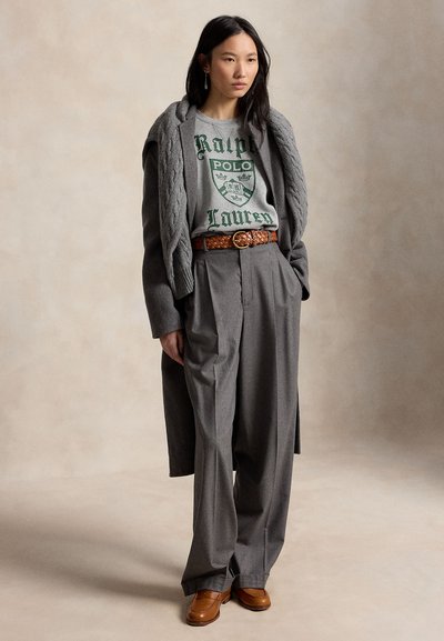 Γυναίκα που φορά γκρι πουλόβερ Ralph Lauren, γκρι φαρδιά παντελόνια, γκρι παλτό, καφέ πλεκτή ζώνη και καφέ μοκασίνια, στέκεται σε ουδέτερο φόντο.