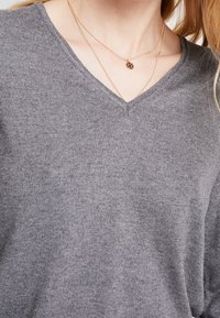 Šedý svetr s V-výstřihem z jemné pleteniny, s nenápadnou texturou. Dva vrstvené zlaté náhrdelníky s kulatými přívěsky doplňují outfit.