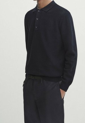 Uomo che indossa una polo nera a manica lunga con texture e bottoni, e pantaloni neri, in piedi con una mano in tasca.