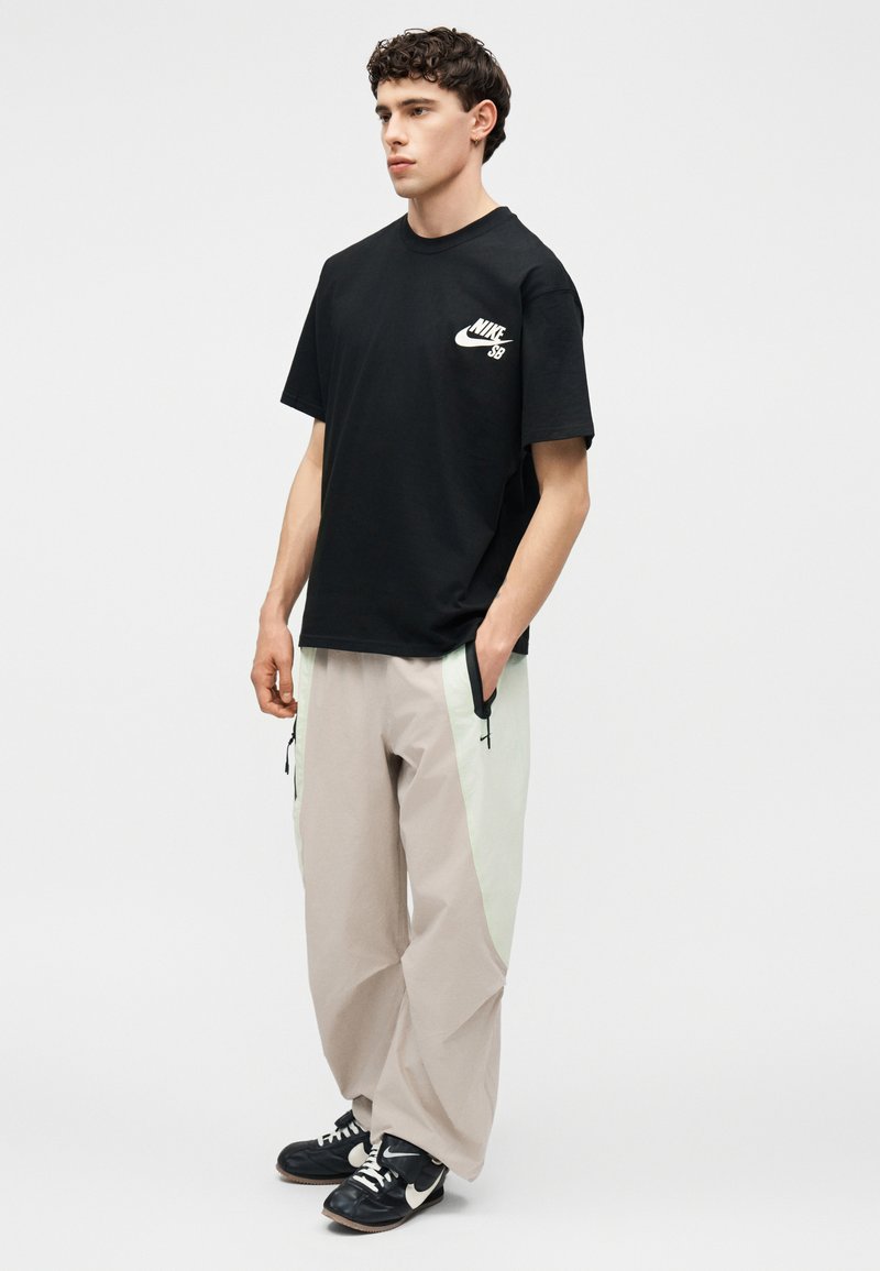 Jonge man staat met handen in de zakken, draagt een zwarte Nike SB t-shirt, beige en witte wijde broek en zwarte Nike sneakers.