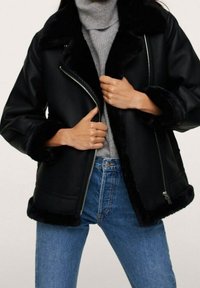 Veste en cuir synthétique noire avec un col et des poignets en shearling. Fermeture éclair, coupe décontractée, associée à un pull col roulé gris et à un jean bleu.