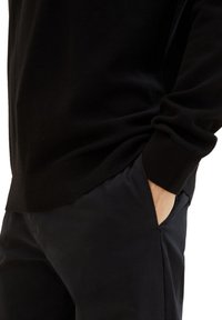 Schwarzer Strickpullover mit gerippten Bündchen, kombiniert mit dunklen Hosen mit Taschen. Der Stoff wirkt weich mit einer strukturierten Oberfläche.