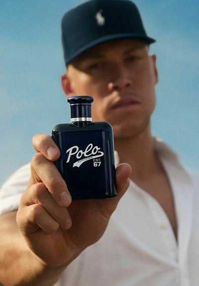 En ung man i en marinblå keps håller en mörkblå Polo doftflaska som visar logotypen, mot en suddig utomhusbakgrund.