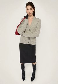 Šedý pletený svetr se zapínáním na pět knoflíků, doplněný černou pouzdrovou sukní a černými kozačkami po kolena. Drží malou vínově červenou kabelku.