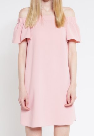 Woman wearing a light pink, off-the-shoulder dress with ruffled sleeves, standing against a plain white background.