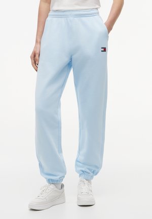 Person trägt hellblaue Jogginghosen mit elastischen Bündchen, weiße Sneaker und ein kleines Tommy Jeans-Logo am linken Oberschenkel.