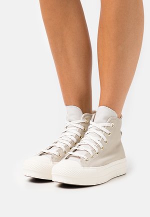 Benedenbenen met lichtbeige hoge sneakers met witte veters en dikke witte rubberen zolen, afgebeeld tegen een effen achtergrond.