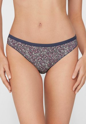 Vrouw draagt laag uitgesneden bikinibroekje met marineblauwe bies en roze-wit bloemenpatroon, staand tegen een effen achtergrond.