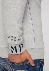 Felpa grigia con polsini a costine, caratterizzata da testi e elementi di design, con una cucitura blu a contrasto. Il materiale sembra morbido e offre una vestibilità informale.