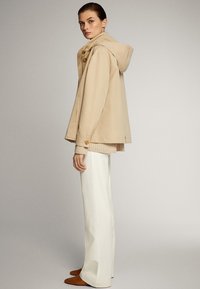 Beige hoodie met houten drukknopen, laag over een lichte gebreide trui, gecombineerd met witte wijde broekspijpen en bruine schoenen.