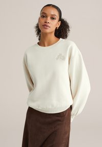 Frau mit lockigem Haar, trägt ein cremefarbenes Sweatshirt mit kleiner Blumenstickerei auf der Brust, kombiniert mit einem dunkelbraunen Rock, steht vor einem schlichten Hintergrund.