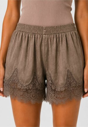 Brune elastiske taljeshorts med bølget blonderkant med blomster, båret af person med solbrun hud og beige ribbet top.