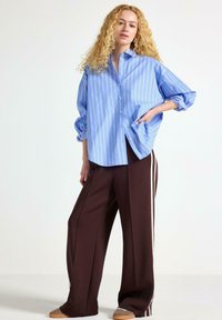 Camicia button-up a righe blu chiaro con maniche arrotolate, abbinata a pantaloni larghi marroni con strisce bianche sui lati. Calzature casual visibili.