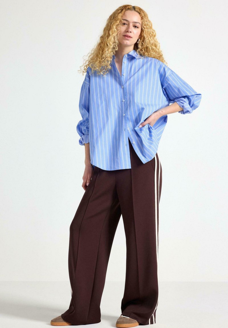 Camicia button-up a righe blu chiaro con maniche arrotolate, abbinata a pantaloni larghi marroni con strisce bianche sui lati. Calzature casual visibili.