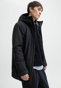 Ung mand iført en sort hættetrøje vinterjakke over en marineblå sweater og en lyseblå skjorte mod en ensfarvet baggrund.