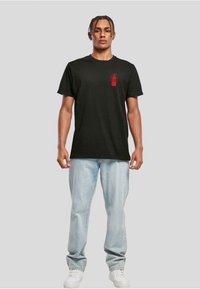 Black cotton t-shirt featuring a red graphic of a fire extinguisher on the left chest area. Model wears light jeans and white sneakers.