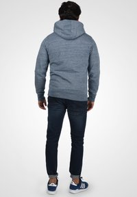 Felpa grigia con finitura texturizzata, dotata di un grande cappuccio, polsini a coste e una vestibilità comoda, abbinata a jeans blu scuro e sneakers blu.