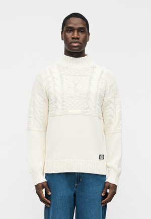 Άνδρας που φοράει λευκό πουλόβερ πλεκτό με υφή και ψηλό γιακά, μαζί με μπλε τζιν, στέκεται μπροστά σε απλό ανοιχτό γκρι φόντο.