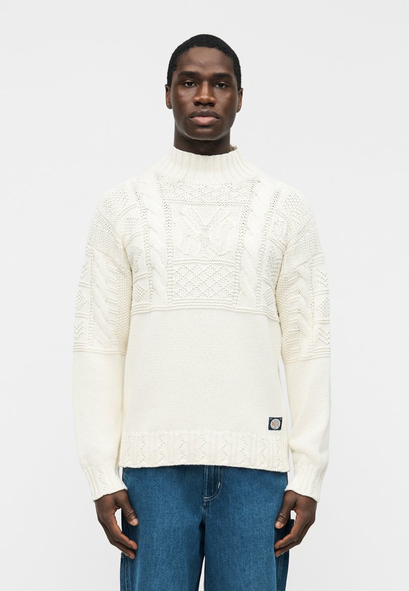 Άνδρας που φοράει λευκό πουλόβερ πλεκτό με υφή και ψηλό γιακά, μαζί με μπλε τζιν, στέκεται μπροστά σε απλό ανοιχτό γκρι φόντο.