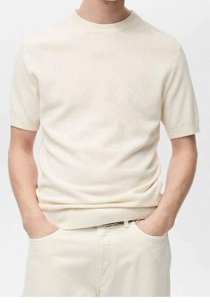 Mand iført en kortærmet, cremefarvet strikket sweater indstukket i lyse beige bukser med hænderne i lommerne mod en ensfarvet baggrund.