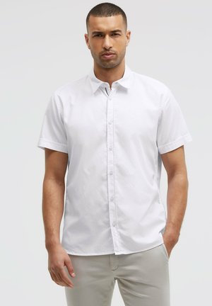 Mann mit kurzen Haaren, der ein weißes kurzärmeliges Hemd und helle Hosen trägt, steht mit einer Hand in der Tasche.