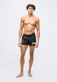 Joven de pie, descalzo y sin camisa, vistiendo pantalones cortos de baño negros con el logo blanco "DSQ" en el muslo izquierdo, sobre un fondo liso de color gris claro.