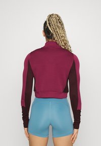 Chaqueta deportiva corta de color granate con espalda texturizada, mangas largas y puños acanalados, combinada con pantalones cortos ajustados de color azul claro.