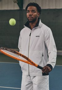 Fehér sportdzseki fekete részletekkel és logóval, hozzá illő rövidnadrággal viselve. Narancssárga teniszütő, amelyet egy levegőben lévő teniszlabda mellett tartanak.