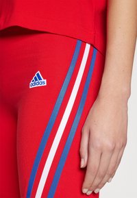 Gros plan sur une main reposant à côté de leggings rouges Adidas présentant des bandes latérales bleues et blanches, ainsi qu'une partie d'un haut rouge assorti.