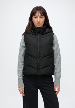 Femme portant une doudoune noire sans manches par-dessus un pull gris à manches longues et un pantalon noir, debout devant un fond blanc uni.