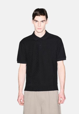 Ragazzo che indossa una polo nera a maniche corte con trama e pantaloni beige, in piedi contro uno sfondo chiaro e uniforme.