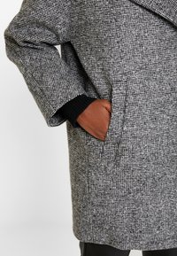 Grijs houndstooth jas met een losse pasvorm, zijzakken en een zwarte geribbelde manchette. De textuur lijkt zacht en geweven.