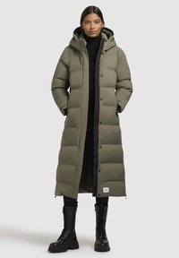 Lang gevoerd parka in olijfgroen met een capuchon, knopen aan de voorkant, zijzakken en een zwarte voering, gecombineerd met zwarte combatboots.