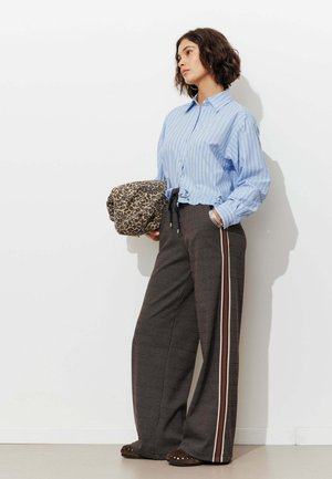 Femme debout contre un mur blanc, portant une chemise rayée bleue, un pantalon large à motifs avec des bandes latérales, tenant un sac imprimé léopard.