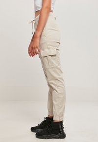 Pantalons cargo beige avec poches latérales, poignets élastiques et taille à cordon, associés à des bottes noires à chevilles. Texture de tissu lisse.