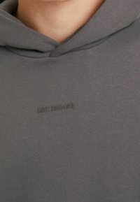 Felpa grigia con cappuccio realizzata in tessuto morbido. Presenta un logo testurizzato in una tonalità più scura, dettagli di cucitura e una vestibilità comoda.