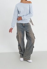 Person som bär en ljusblå avskuren sweatshirt, vida blekta cargobyxor med flera fickor och vita sneakers, stående mot en vit bakgrund.