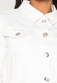 Biała denimowa kurtka z wyraźnym kołnierzykiem, srebrnymi zatrzaskami i kieszeniami z przodu. Tkanina ma gładką fakturę z widocznymi detalami szycia.