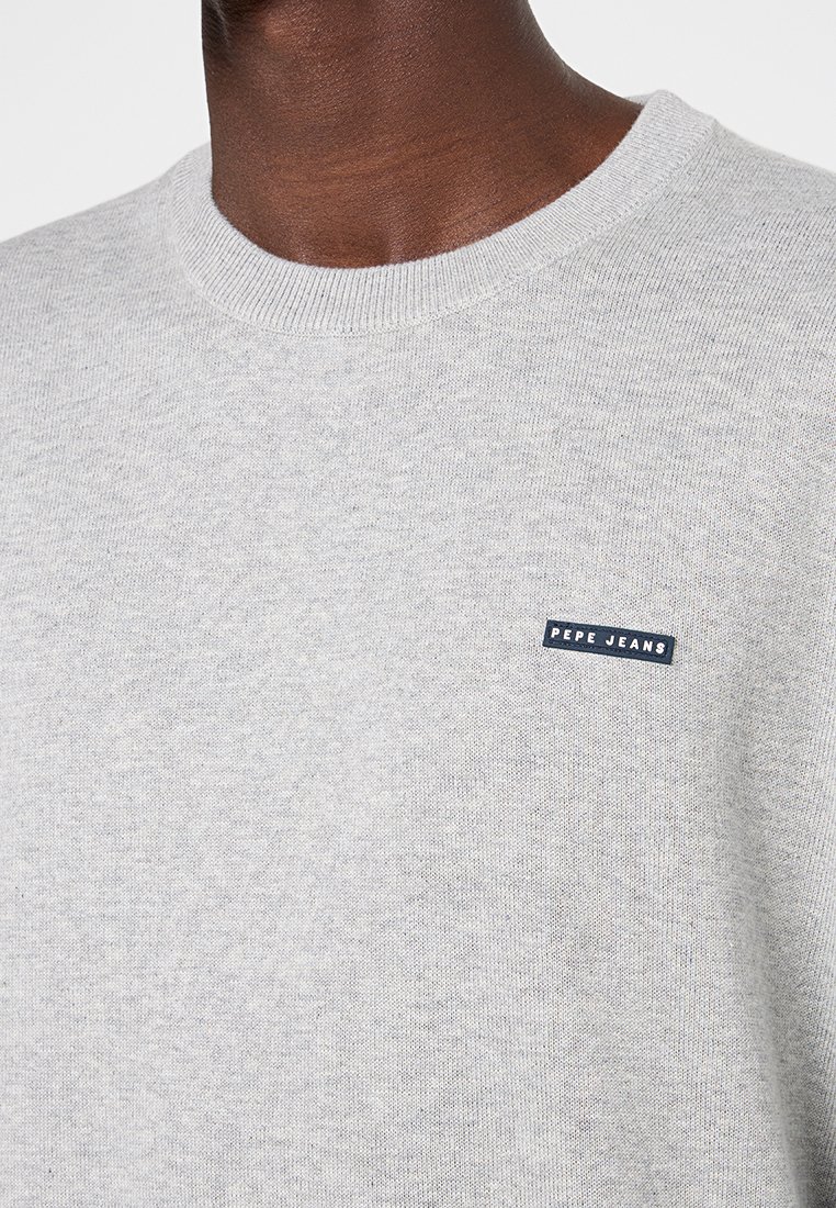 Gros plan sur une personne à la peau foncée portant un pull léger gris de Pepe Jeans avec un col rond et un petit patch logo noir sur la poitrine.