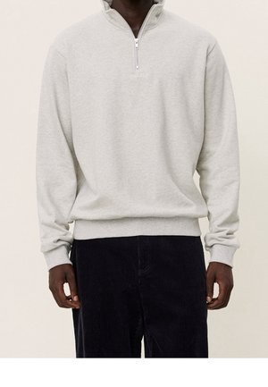 Man draagt een lichtgrijze pullover met kwartritssluiting, geribde boorden en taille, gecombineerd met een donkere ribfluwelen broek.