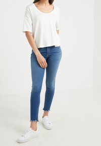 Top blanco de manga corta y escote redondo combinado con jeans skinny azules de cintura media con dobladillos deshilachados y zapatillas blancas.