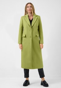 Long, collared coat in bright green wool blend, featuring two front pockets and two matching buttons. Worn over a black top and leather pants.