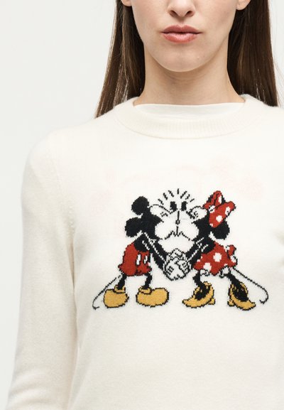 Γυναίκα που φοράει λευκό πουλόβερ με πλεκτό σχέδιο δύο καρτούν ποντικιών που κρατιούνται χέρι-χέρι και φιλιούνται, το ένα φοράει κόκκινα σορτς και το άλλο ένα κόκκινο φόρεμα.