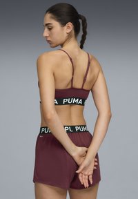 Marron sportski grudnjak s tankim naramenicama, s crnim pojasom na kojem se nalazi "PUMA" logo; uparen s odgovarajućim marron kratkim hlačama, glatke teksture.