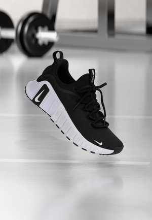 Schwarz-weißer Nike Free Laufschuh mit dicker Sohle, der über einem Fitnessboden schwebt, Hantel und Gestell im unscharfen Hintergrund.