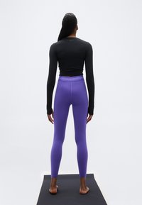 Άτομο που στέκεται ξυπόλυτο σε μαύρο στρώμα γιόγκα, φορώντας μαύρο μακρυμάνικο μπλουζάκι και μοβ κολάν, γυρισμένο πλάτη σε λευκό φόντο.