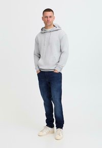 Sudadera con capucha gris y bolsillo frontal, combinada con jeans oscuros y zapatillas blancas. Diseño simple, material de algodón, corte relajado.