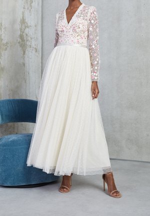 Kobieta w długiej białej sukni z przezroczystymi rękawami i górą ozdobionymi kwiatowym haftem, stojąca obok niebieskiego tapicerowanego fotela.