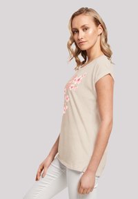 Beige Baumwoll-T-Shirt mit kurzen Ärmeln, mit pinkem Blumenmuster und einem weiten Rundhalsausschnitt. Kombiniert mit weißen Hosen.