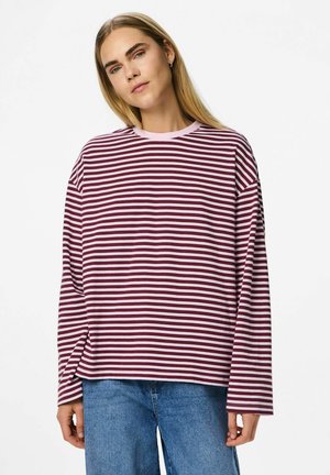 Ung kvinde iført en langærmet marsala- og hvidstribet skjorte samt blå jeans, stående mod en ensfarvet hvid baggrund.